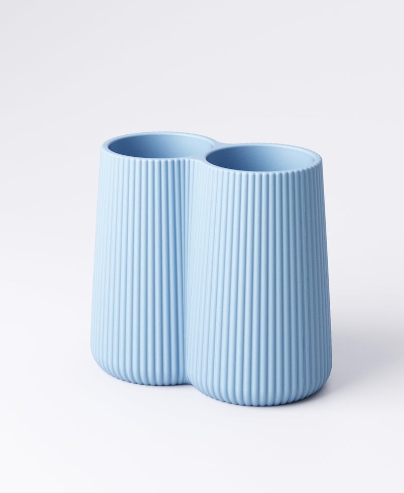 May include: A light blue, double-chambered vase with vertical ribbed detailing. The vase has a rounded base and a smooth rim. The two chambers are connected, creating a unique, modern design. The vase is made of a solid material and is suitable for home decor.