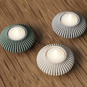 Puede incluir: Tres portavelas para velas de t&eacute; con un dise&ntilde;o circular y acanalado. Cada soporte tiene una vela blanca con una llama parpadeante. Los soportes son en tonos de verde, gris y blanco, colocados sobre una superficie de madera.