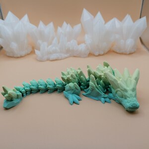K&ouml;nnte beinhalten: Ein 3D-gedrucktes, bewegliches Drachenspielzeug in T&uuml;rkis- und Gr&uuml;nt&ouml;nen mit kristallartigem Design. Im Hintergrund befinden sich mehrere wei&szlig;e Kristallformationen. Der Drache liegt auf einer hellbraunen Oberfl&auml;che.