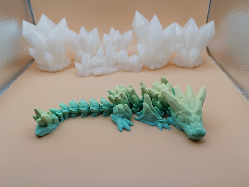 K&ouml;nnte beinhalten: Ein 3D-gedrucktes, bewegliches Drachenspielzeug in T&uuml;rkis- und Gr&uuml;nt&ouml;nen mit kristallartigem Design. Im Hintergrund befinden sich mehrere wei&szlig;e Kristallformationen. Der Drache liegt auf einer hellbraunen Oberfl&auml;che.