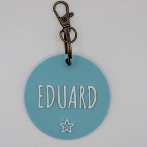 May include: A round, light blue keychain tag with the name "EDUARD" in white, outlined letters. A small white star is below the name. The tag is attached to a bronze-colored metal clasp.