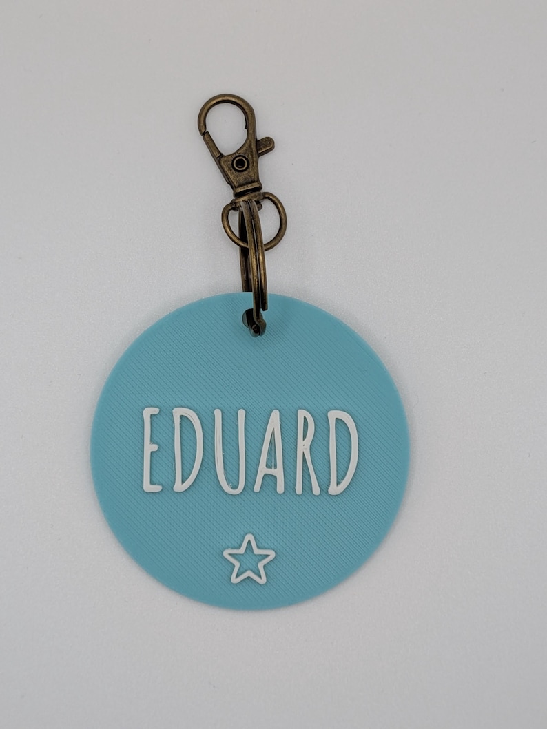 May include: A round, light blue keychain tag with the name "EDUARD" in white, outlined letters. A small white star is below the name. The tag is attached to a bronze-colored metal clasp.
