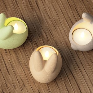 K&ouml;nnte beinhalten: Drei hasenf&ouml;rmige Kerzenhalter in Pastellfarben: Hellgr&uuml;n, Beige und Grau. Jeder Halter enth&auml;lt eine brennende, runde Kerze. Die Halter bestehen aus einem strukturierten Material und stehen auf einer Holzoberfl&auml;che. Die Kerzen erzeugen ein warmes Leuchten.