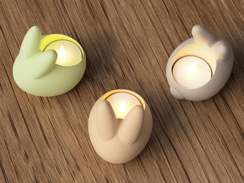 K&ouml;nnte beinhalten: Drei hasenf&ouml;rmige Kerzenhalter in Pastellfarben: Hellgr&uuml;n, Beige und Grau. Jeder Halter enth&auml;lt eine brennende, runde Kerze. Die Halter bestehen aus einem strukturierten Material und stehen auf einer Holzoberfl&auml;che. Die Kerzen erzeugen ein warmes Leuchten.