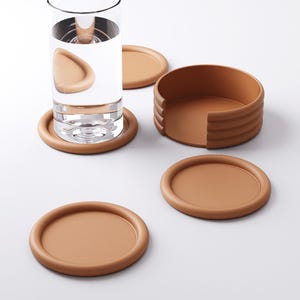 Puede incluir: Un juego de posavasos marrones con un soporte a juego. Un posavasos est&aacute; debajo de un vaso de agua. Los posavasos son redondos con un borde elevado. El soporte es un recipiente marr&oacute;n semicircular con un dise&ntilde;o acanalado.