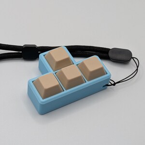 May include: A light blue keyboard keycap holder with four beige keycaps. The holder is connected to a black lanyard. The keycaps are arranged in a T-shape. The background is a solid light gray.