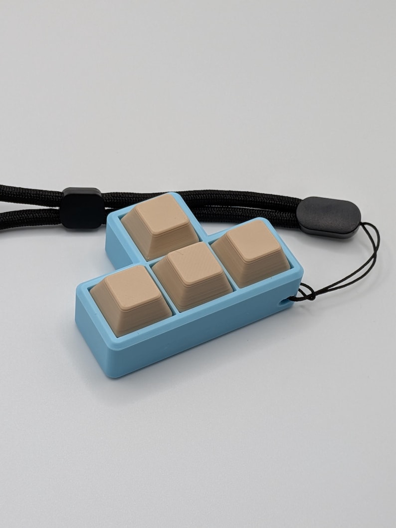 May include: A light blue keyboard keycap holder with four beige keycaps. The holder is connected to a black lanyard. The keycaps are arranged in a T-shape. The background is a solid light gray.
