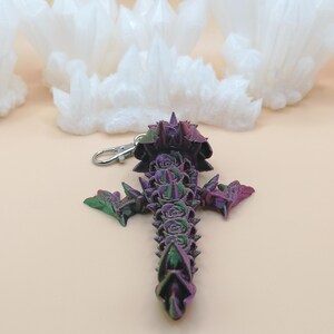 May include: A purple, green, and pink cross-shaped keychain with a silver clasp. The cross is decorated with rose-like details and pointed edges. The background features white, cloud-like shapes.