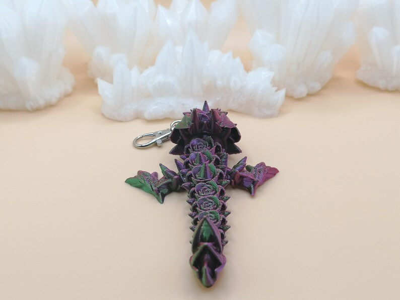 May include: A purple, green, and pink cross-shaped keychain with a silver clasp. The cross is decorated with rose-like details and pointed edges. The background features white, cloud-like shapes.