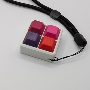 May include: A white square keychain with four colorful keycaps in shades of red, pink, and purple. The keychain has a black lanyard attached. The keycaps are arranged in a 2x2 grid.