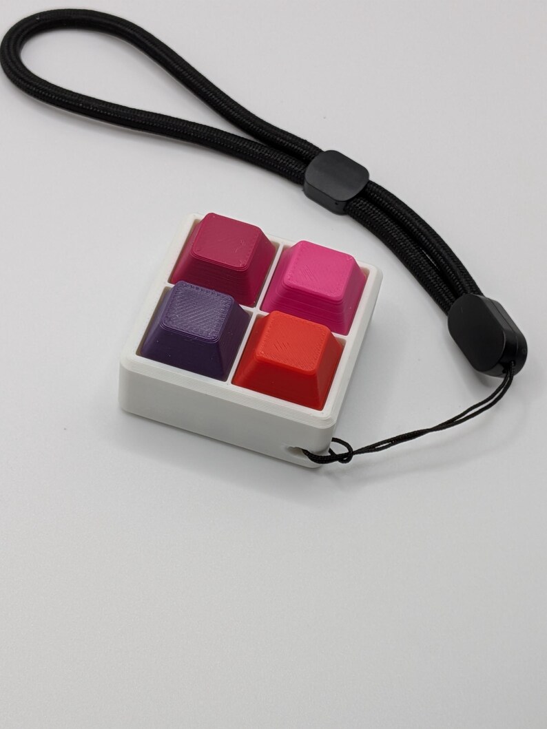 May include: A white square keychain with four colorful keycaps in shades of red, pink, and purple. The keychain has a black lanyard attached. The keycaps are arranged in a 2x2 grid.