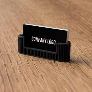 May include: A black business card holder with a rectangular design, holding a stack of black business cards. The cards display the text "COMPANY LOGO" in white. The holder is on a wooden surface.