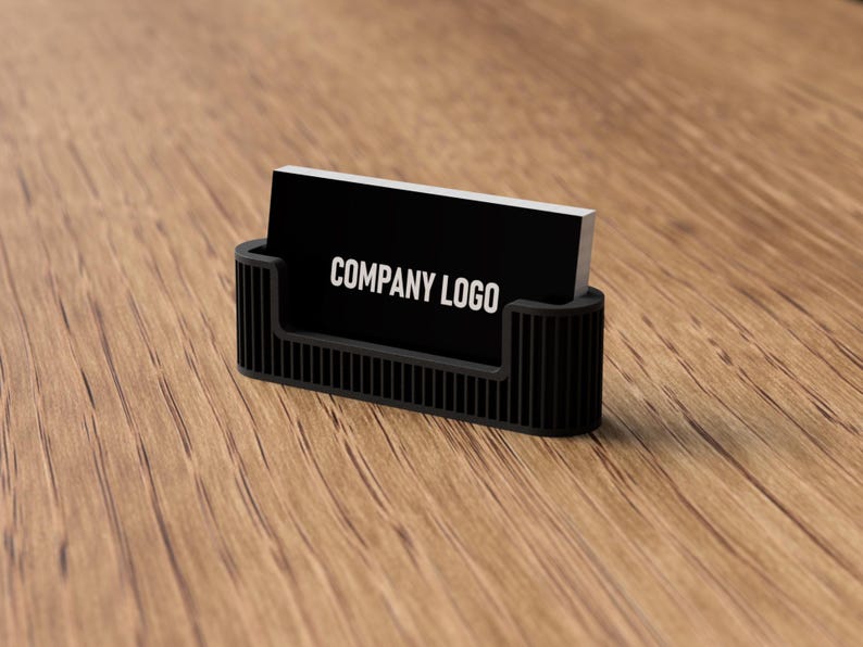 May include: A black business card holder with a rectangular design, holding a stack of black business cards. The cards display the text "COMPANY LOGO" in white. The holder is on a wooden surface.