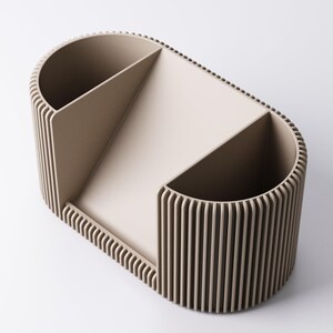 May include: A beige desk organiser with a distinctive design. The organiser has a rounded rectangular shape with vertical ribbed sides and interior dividers. It features two open compartments and a sloped section for storage.