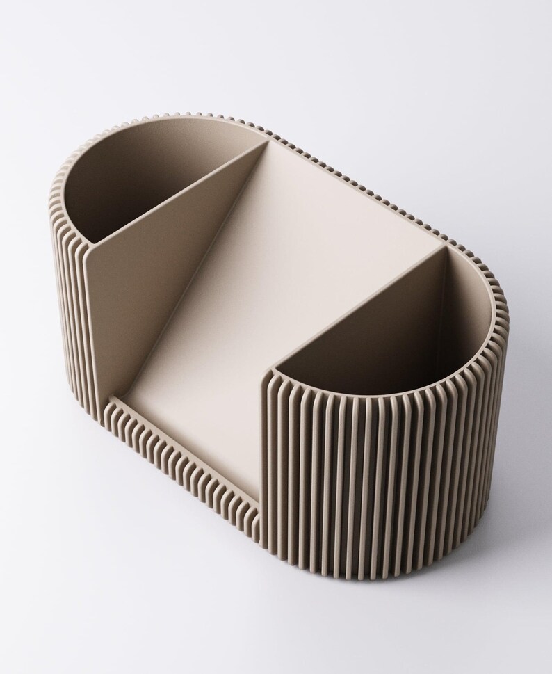 May include: A beige desk organiser with a distinctive design. The organiser has a rounded rectangular shape with vertical ribbed sides and interior dividers. It features two open compartments and a sloped section for storage.