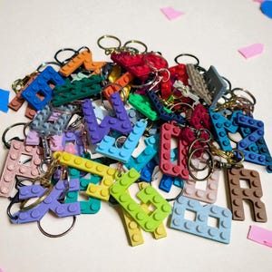 May include: A collection of colorful letter-shaped keychains, resembling building blocks. The keychains are in various colors, including blue, green, yellow, and red, with gold-colored key rings attached. The letters are scattered on a light background.