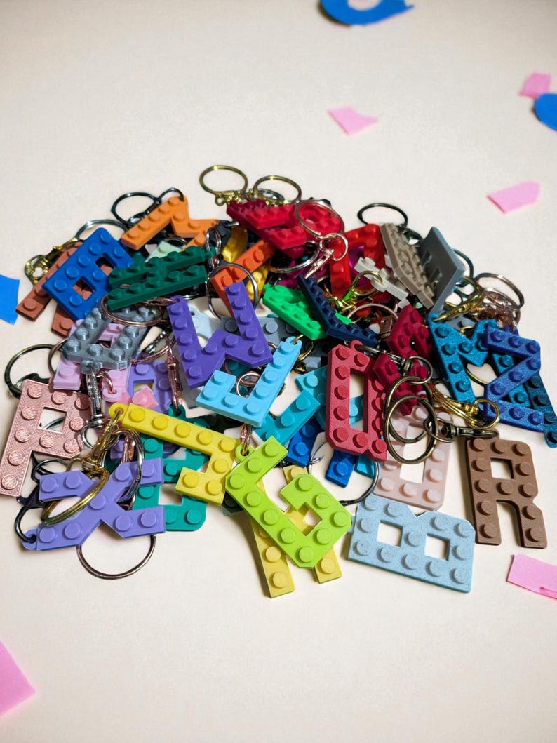 May include: A collection of colorful letter-shaped keychains, resembling building blocks. The keychains are in various colors, including blue, green, yellow, and red, with gold-colored key rings attached. The letters are scattered on a light background.