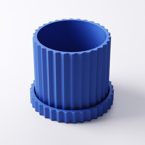 May include: A cobalt blue cylindrical planter with a matching saucer. The planter has vertical, ridged detailing around the exterior and a zig-zag pattern on the rim and saucer edge. The planter is designed to hold plants.