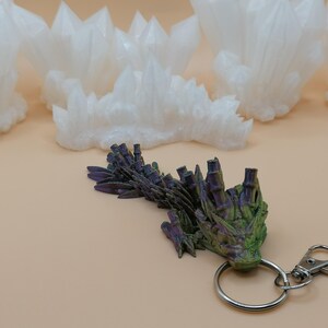 May include: A green, purple, and black dragon keychain with a silver clasp and ring. Several white crystal formations are in the background, all on a tan surface.