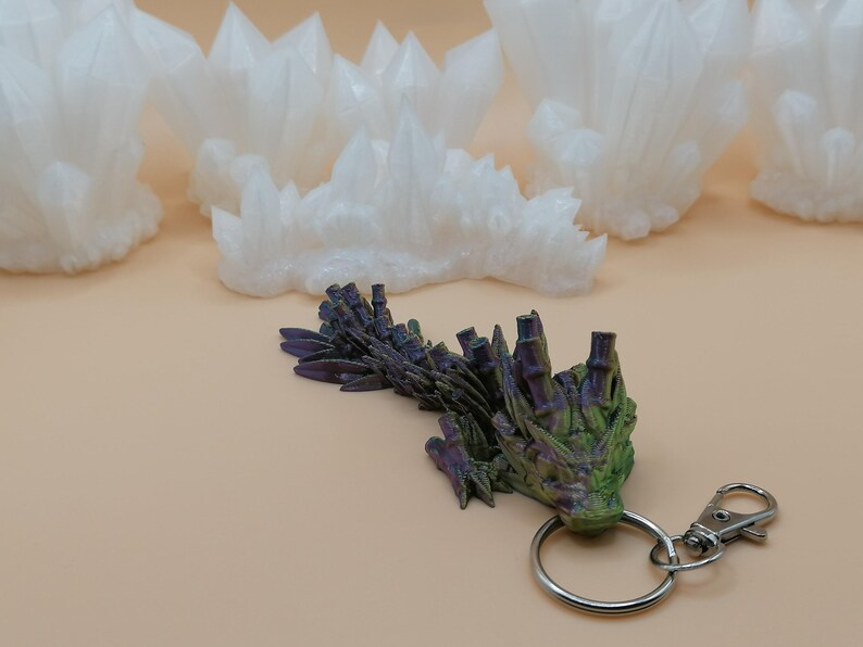 May include: A green, purple, and black dragon keychain with a silver clasp and ring. Several white crystal formations are in the background, all on a tan surface.