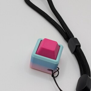 May include: A 3D-printed keycap with a pink top and a gradient blue-pink base, attached to a black wrist strap. The keycap is square-shaped and has a textured surface. The wrist strap is made of woven black material.