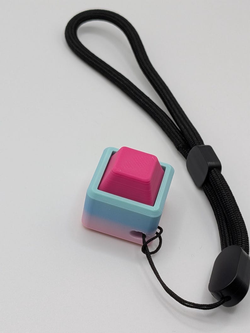 May include: A 3D-printed keycap with a pink top and a gradient blue-pink base, attached to a black wrist strap. The keycap is square-shaped and has a textured surface. The wrist strap is made of woven black material.