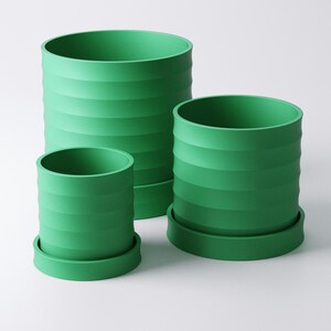 Op de afbeelding: Drie groene cilindrische bloempotten van verschillende maten. Elke pot heeft een geribbeld ontwerp en staat op een bijpassende ronde basis. De potten zijn felgroen van kleur en staan tegen een witte achtergrond.