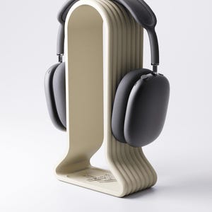 May include: A pair of black over-ear headphones resting on a beige headphone stand. The stand has a curved design with vertical ridges and a small tray at the base. The headphones have a gray headband and black earcups. The stand is on a white background.