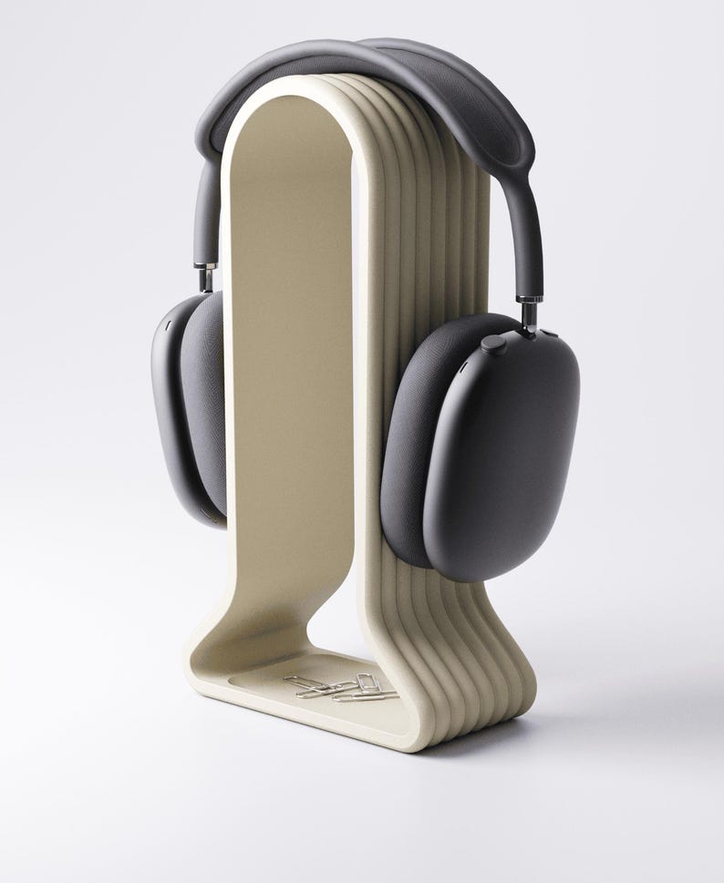 May include: A pair of black over-ear headphones resting on a beige headphone stand. The stand has a curved design with vertical ridges and a small tray at the base. The headphones have a gray headband and black earcups. The stand is on a white background.