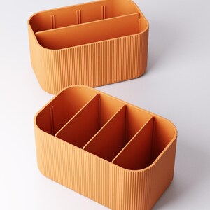 May include: Two orange storage organisers with vertical ribbed sides and internal dividers. The organisers have rounded corners and are designed to hold various items. The dividers create multiple compartments for organisation. The colour is a warm, inviting orange.