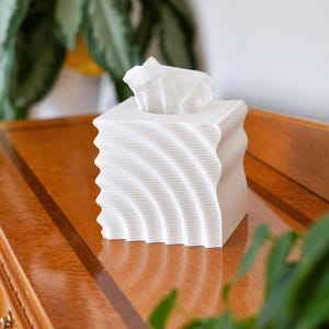 May include: A square white tissue box cover featuring a distinct rippled, wave-like texture on all four sides. The top surface has a central rectangular opening through which a single white facial tissue is pulled upward and visible.