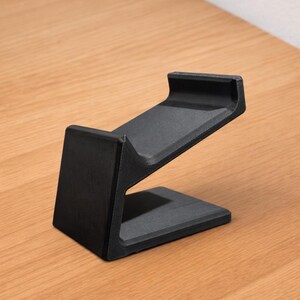 May include: A black, angled stand, likely for a phone or tablet, is displayed on a wooden surface. The stand has a rectangular base and a sloped top section with a lip to hold a device. The design is minimalist and modern.