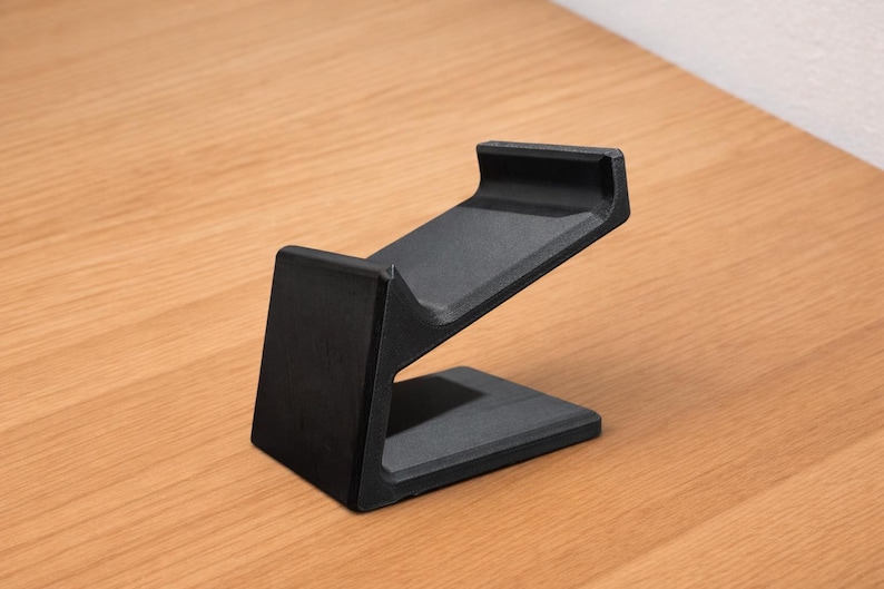 May include: A black, angled stand, likely for a phone or tablet, is displayed on a wooden surface. The stand has a rectangular base and a sloped top section with a lip to hold a device. The design is minimalist and modern.