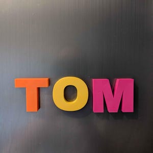 May include: The name "TOM" is spelled out in colorful, three-dimensional letters. The "T" is orange, the "O" is yellow, and the "M" is pink. The letters are placed against a dark gray, textured background.