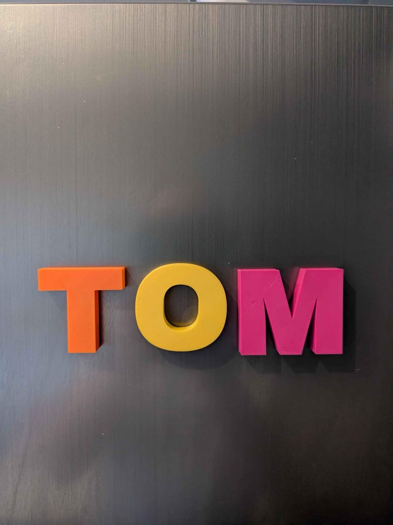 May include: The name "TOM" is spelled out in colorful, three-dimensional letters. The "T" is orange, the "O" is yellow, and the "M" is pink. The letters are placed against a dark gray, textured background.