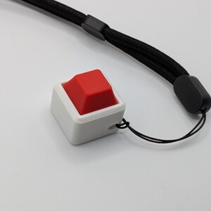 May include: A white square-shaped button with a red top, attached to a black wrist strap. The button has a small hole for the string. The wrist strap is made of a woven material.