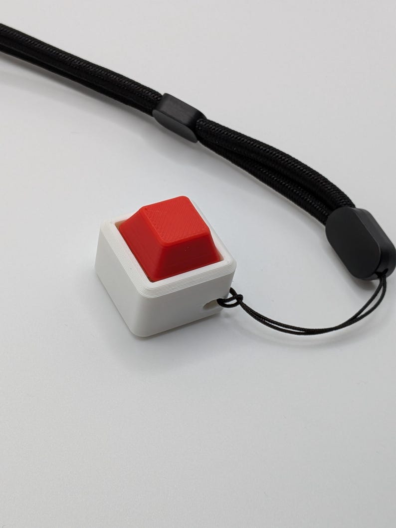 May include: A white square-shaped button with a red top, attached to a black wrist strap. The button has a small hole for the string. The wrist strap is made of a woven material.