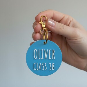May include: A light blue circular keychain tag with the name "OLIVER" and "CLASS 3B" in white lettering. The tag is attached to a gold-colored clasp and ring.