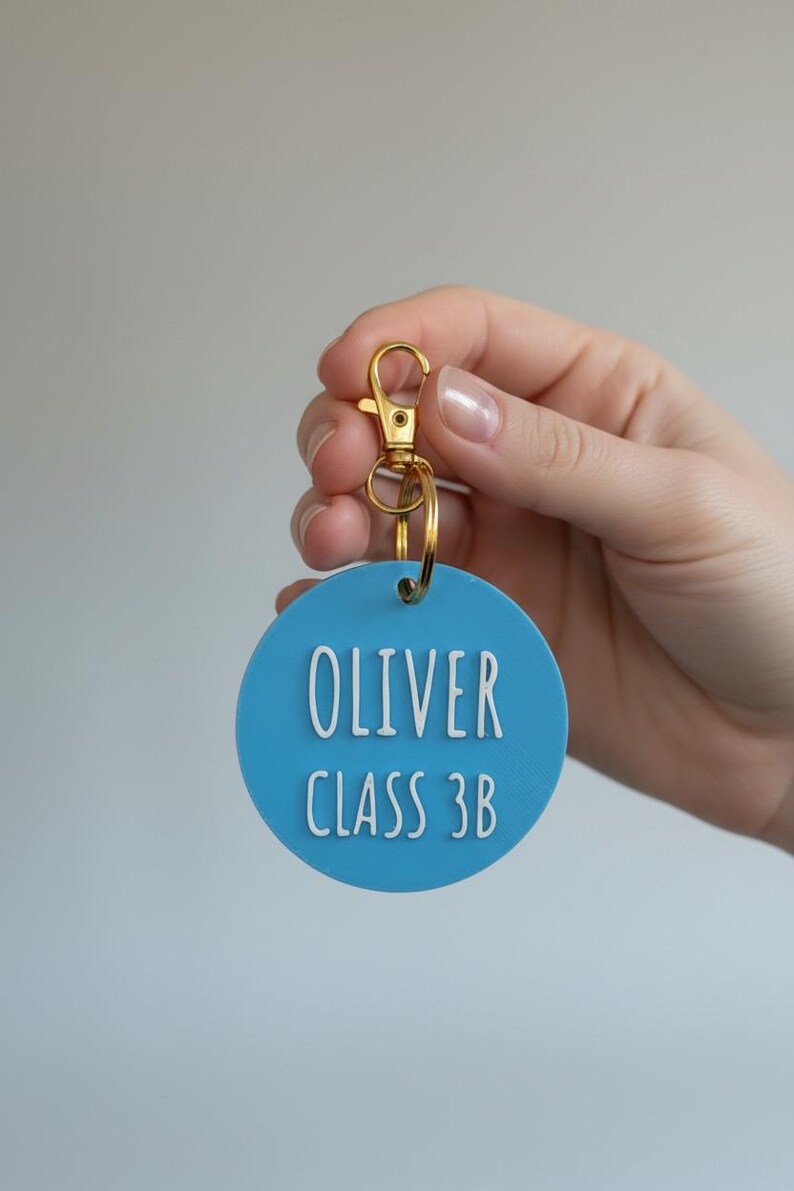 May include: A light blue circular keychain tag with the name "OLIVER" and "CLASS 3B" in white lettering. The tag is attached to a gold-colored clasp and ring.