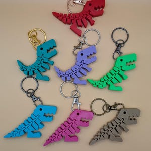 May include: A collection of colorful dinosaur keychains in red, teal, purple, green, blue, pink, and gray. Each keychain features a segmented dinosaur design with a metal keyring and clasp.