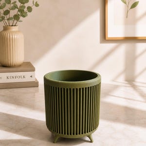 Puede incluir: Jardinera cil&iacute;ndrica verde oliva con dise&ntilde;o de costillas verticales. La jardinera tiene tres patas peque&ntilde;as y est&aacute; sobre una superficie clara. Un jarr&oacute;n beige con ramas de eucalipto y una impresi&oacute;n bot&aacute;nica enmarcada est&aacute;n en el fondo.