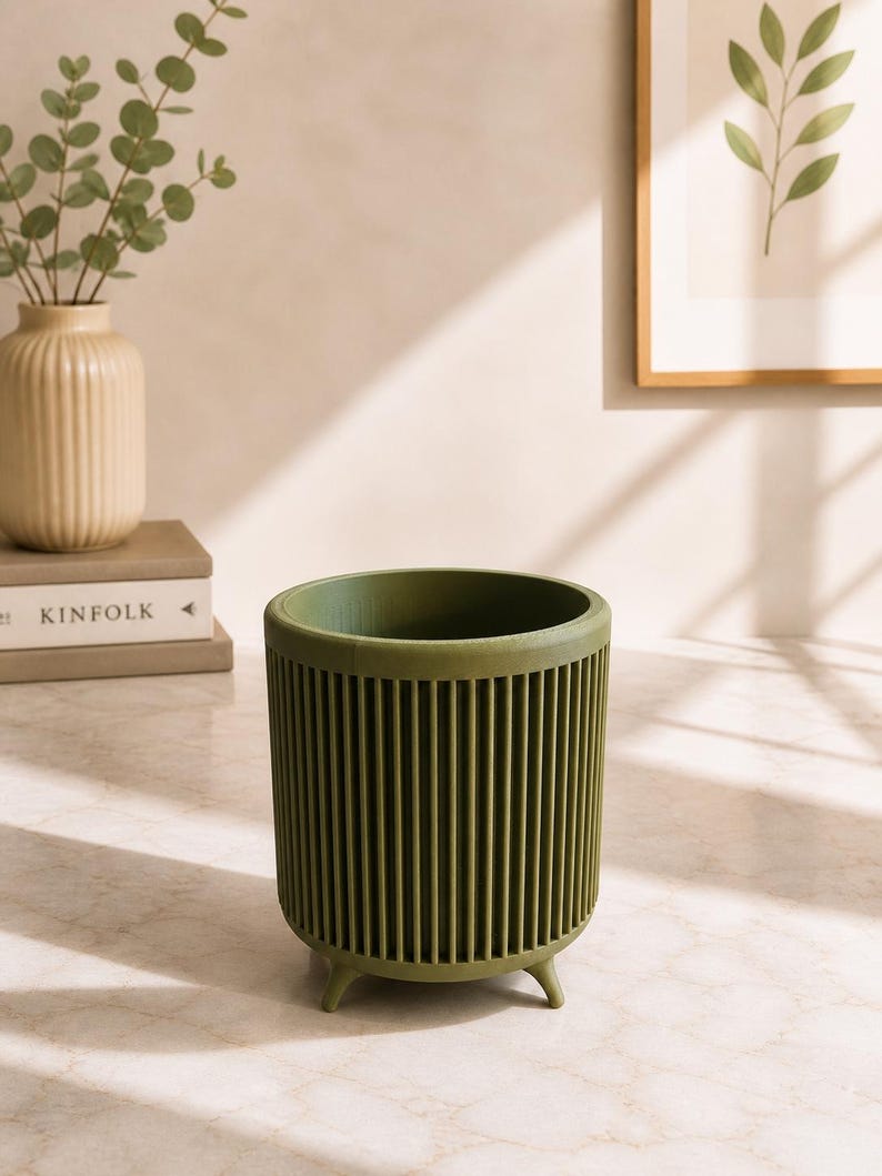 Puede incluir: Jardinera cil&iacute;ndrica verde oliva con dise&ntilde;o de costillas verticales. La jardinera tiene tres patas peque&ntilde;as y est&aacute; sobre una superficie clara. Un jarr&oacute;n beige con ramas de eucalipto y una impresi&oacute;n bot&aacute;nica enmarcada est&aacute;n en el fondo.