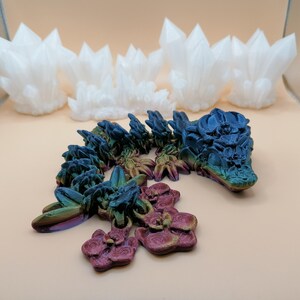 May include: A 3D-printed articulated dragon toy in iridescent colors of blue, green, and purple, with a matching flower-like charm. Several white crystal-shaped sculptures are in the background.