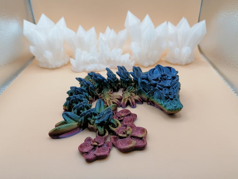 May include: A 3D-printed articulated dragon toy in iridescent colors of blue, green, and purple, with a matching flower-like charm. Several white crystal-shaped sculptures are in the background.