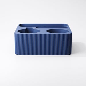 May include: A rectangular blue desk organizer featuring a ribbed exterior texture. The top surface contains multiple recessed compartments of varying shapes, including two circular openings and two elongated slots for holding stationery items.