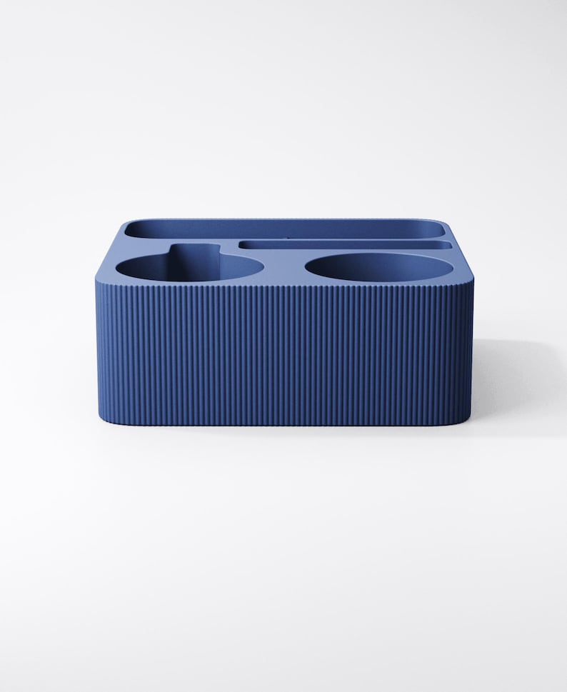 May include: A rectangular blue desk organizer featuring a ribbed exterior texture. The top surface contains multiple recessed compartments of varying shapes, including two circular openings and two elongated slots for holding stationery items.