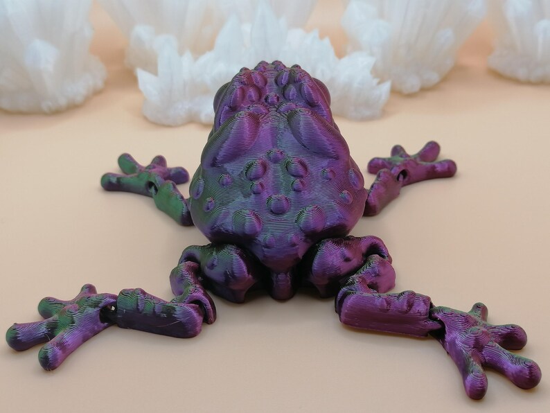 May include: A 3D-printed frog figurine in shades of purple, green, and blue. The frog has a textured body and articulated limbs. The background features white, crystalline formations.