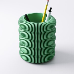 May include: A green, cylindrical desk organizer with a ribbed, wavy design. The organizer holds a yellow pencil, a white marker, and a black pen. The interior of the organizer is a solid green color. The background is white.