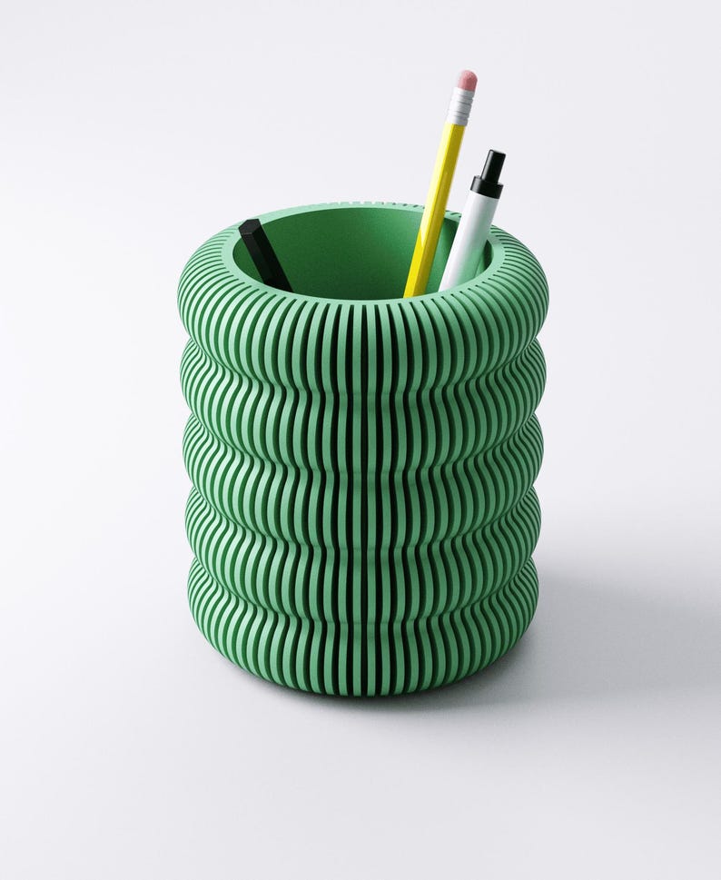 May include: A green, cylindrical desk organizer with a ribbed, wavy design. The organizer holds a yellow pencil, a white marker, and a black pen. The interior of the organizer is a solid green color. The background is white.