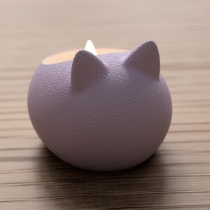 May include: A lavender-colored, cat-shaped candle holder with two pointed ears. The candle holder has a textured surface and contains a lit candle. The background is a light brown wooden surface.
